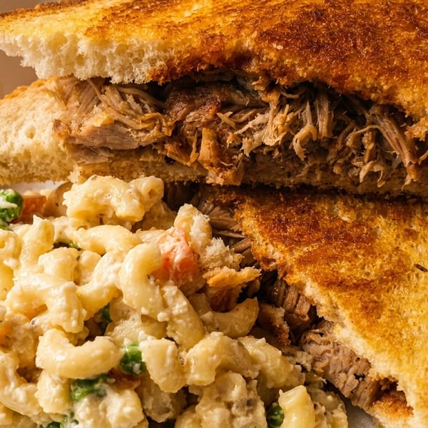 Pulled pork sandwich on toasted bread with a side of macaroni salad in a takeout container.