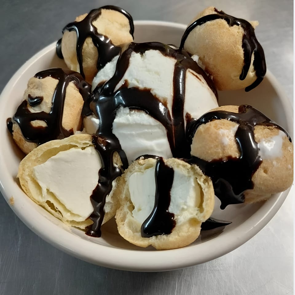 A white bowl filled with cream puffs and vanilla ice cream, drizzled with chocolate syrup.