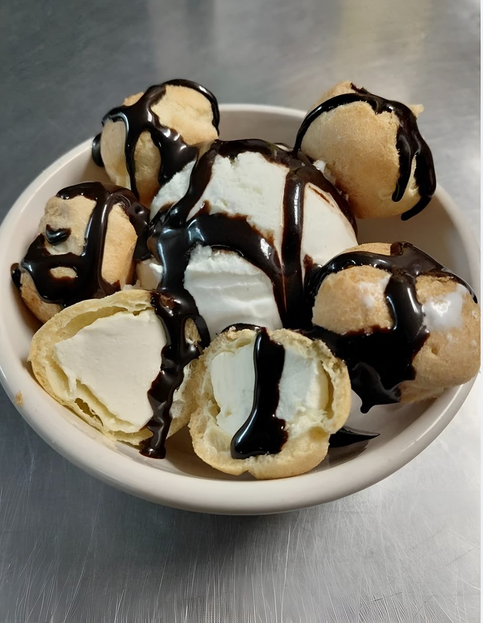 A white bowl filled with cream puffs and vanilla ice cream, drizzled with chocolate syrup.