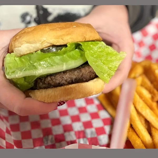 Hands holding a burger with lettuce and pickles, next to a basket of french fries.