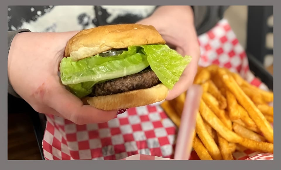 Hands holding a burger with lettuce and pickles, next to a basket of french fries.