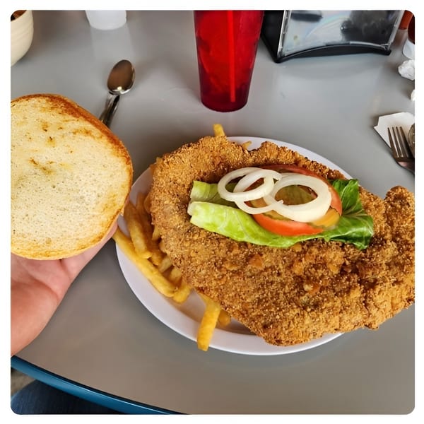 A large fried fish sandwich with lettuce, tomato, and onion, served with fries and a drink.