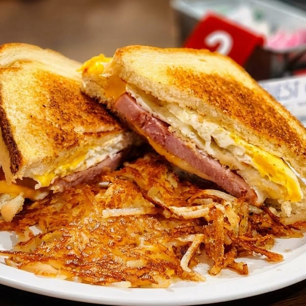 A toasted breakfast sandwich with egg, ham, and cheese, served with crispy hash browns.