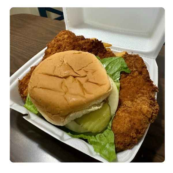 Fried chicken sandwich with lettuce, onion, and pickles, alongside chicken tenders and fries in a takeout container.