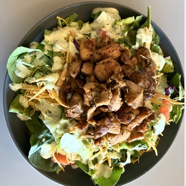 A dark bowl filled with a fresh salad topped with grilled chicken, cheese, and creamy dressing.