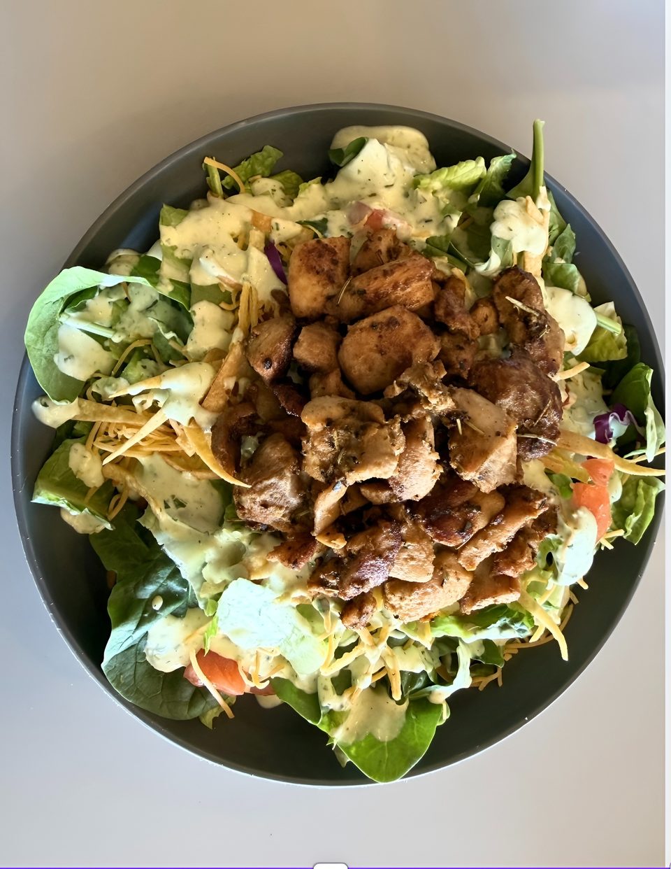 A dark bowl filled with a fresh salad topped with grilled chicken, cheese, and creamy dressing.
